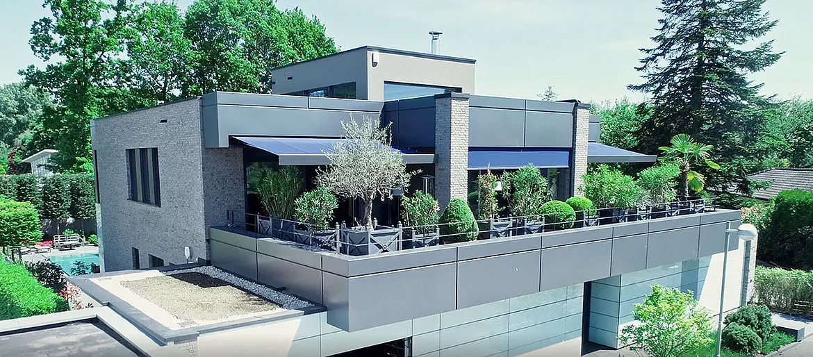 A modern three-story house is photographed sideways from the left with a large, landscaped balcony on the 1st floor and surrounded by other houses and trees. 