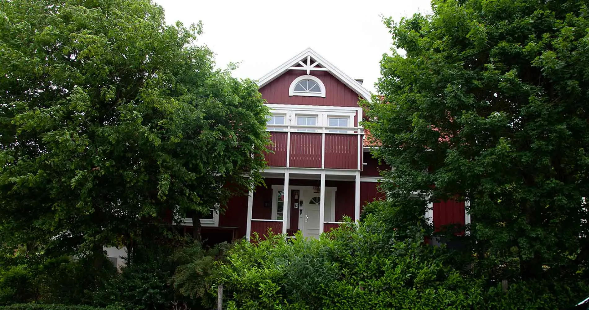A house with its front door, a balcony above it and the gable peeks out between two even larger trees. More bushes can be seen in the foreground.