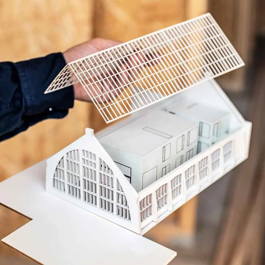 A person lifts the roof from the paper model of the brick house to look at the paper palm house below.