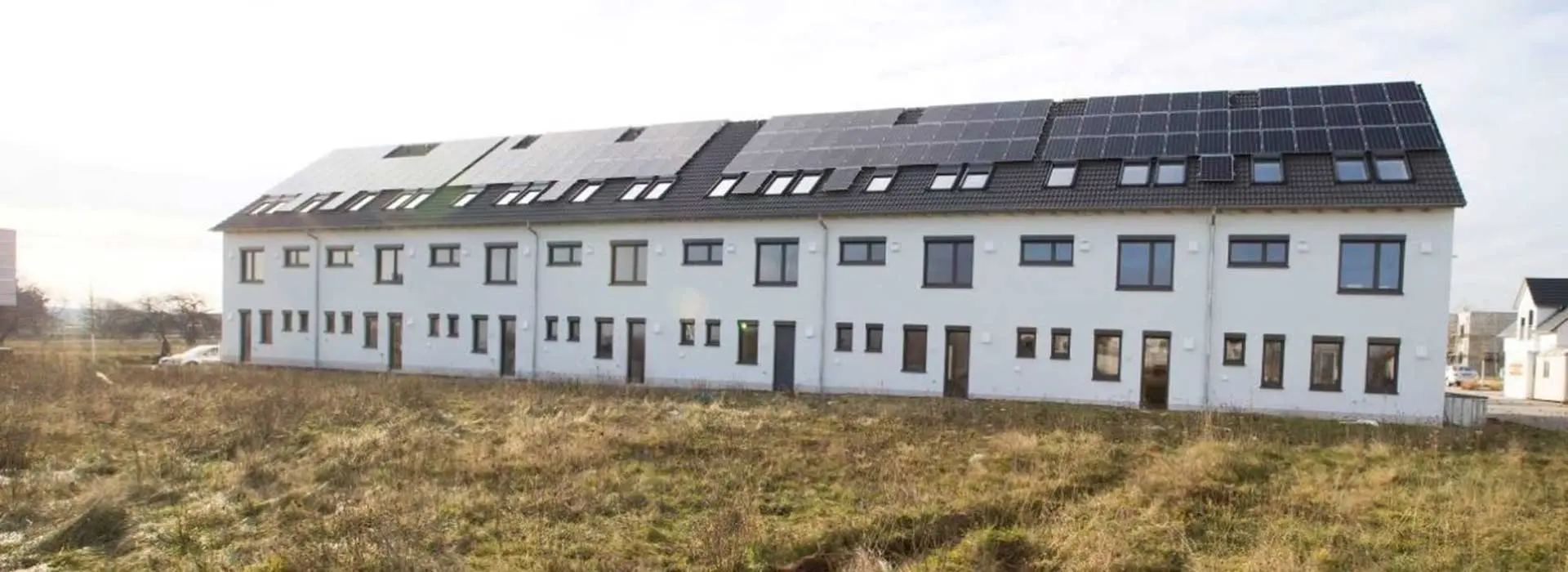 A long row house with a meadow in the foreground and photovoltaics on the roof stands in a housing estate.