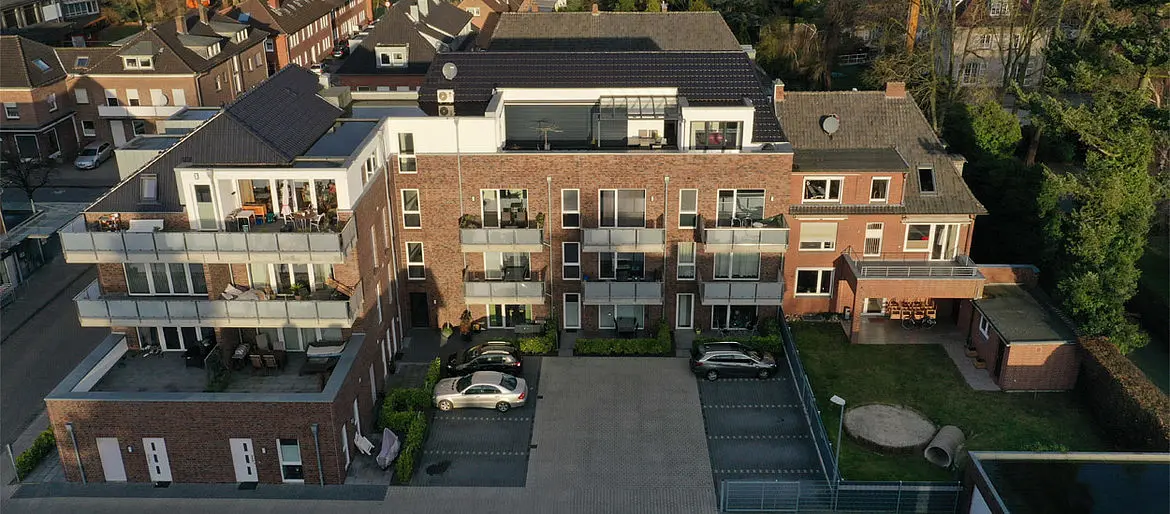 An apartment house in a housing estate has balconies, parking spaces in the foreground and a garden on its right side in front of the house.