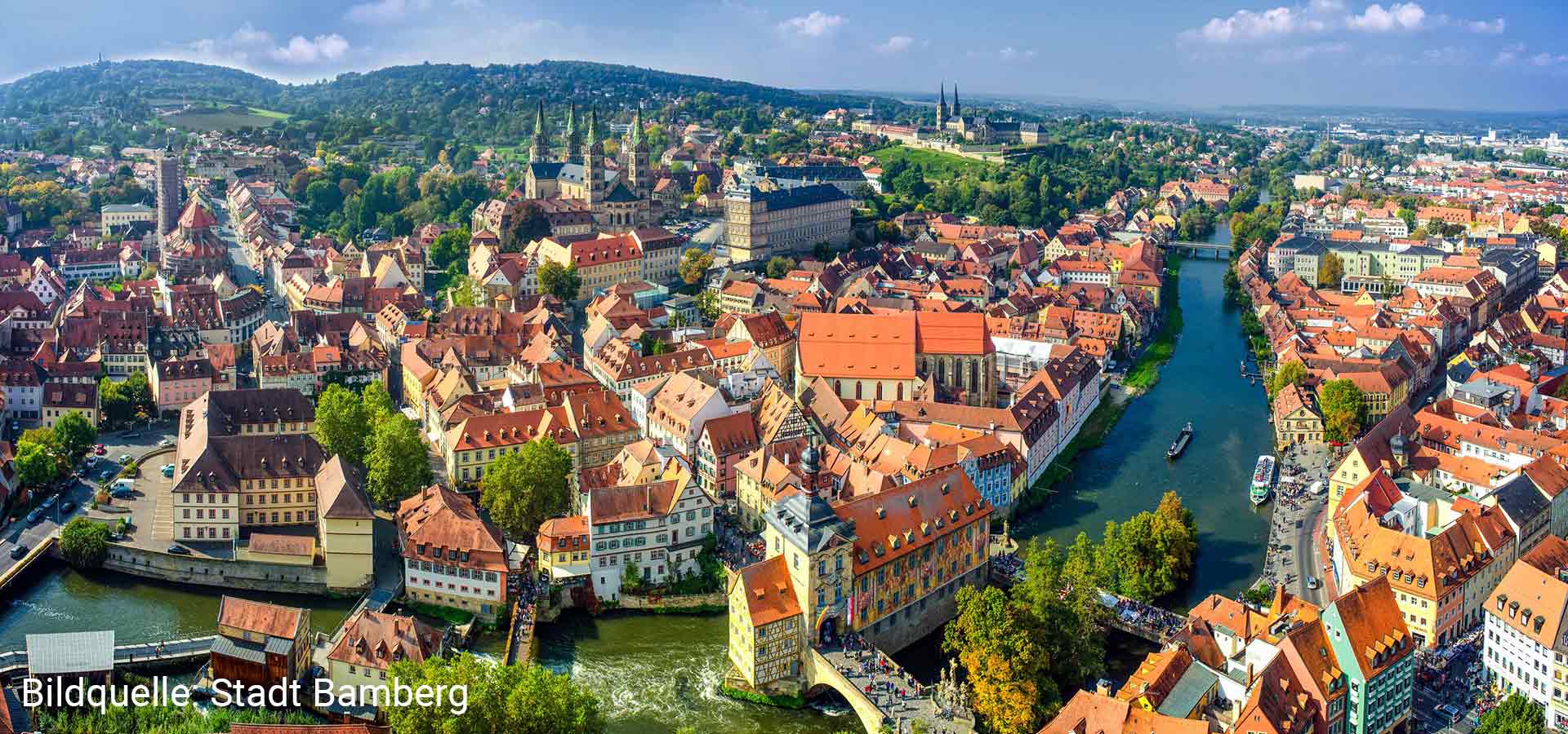 In an aerial view, one looks over the old town of Bamberg with old half-timbered houses built closely together and a river in the foreground that runs under several bridges as well as large churches in the background.