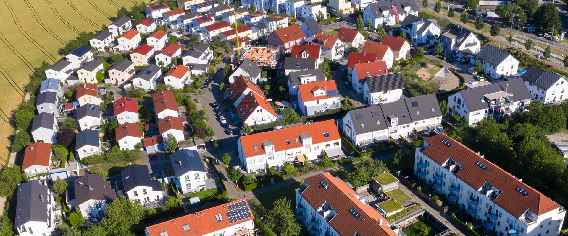 A residential area was photographed from a bird's eye view, with small streets and gardens. In the middle, you can see an unfinished house, next to a crane. In the background, you can see a field.