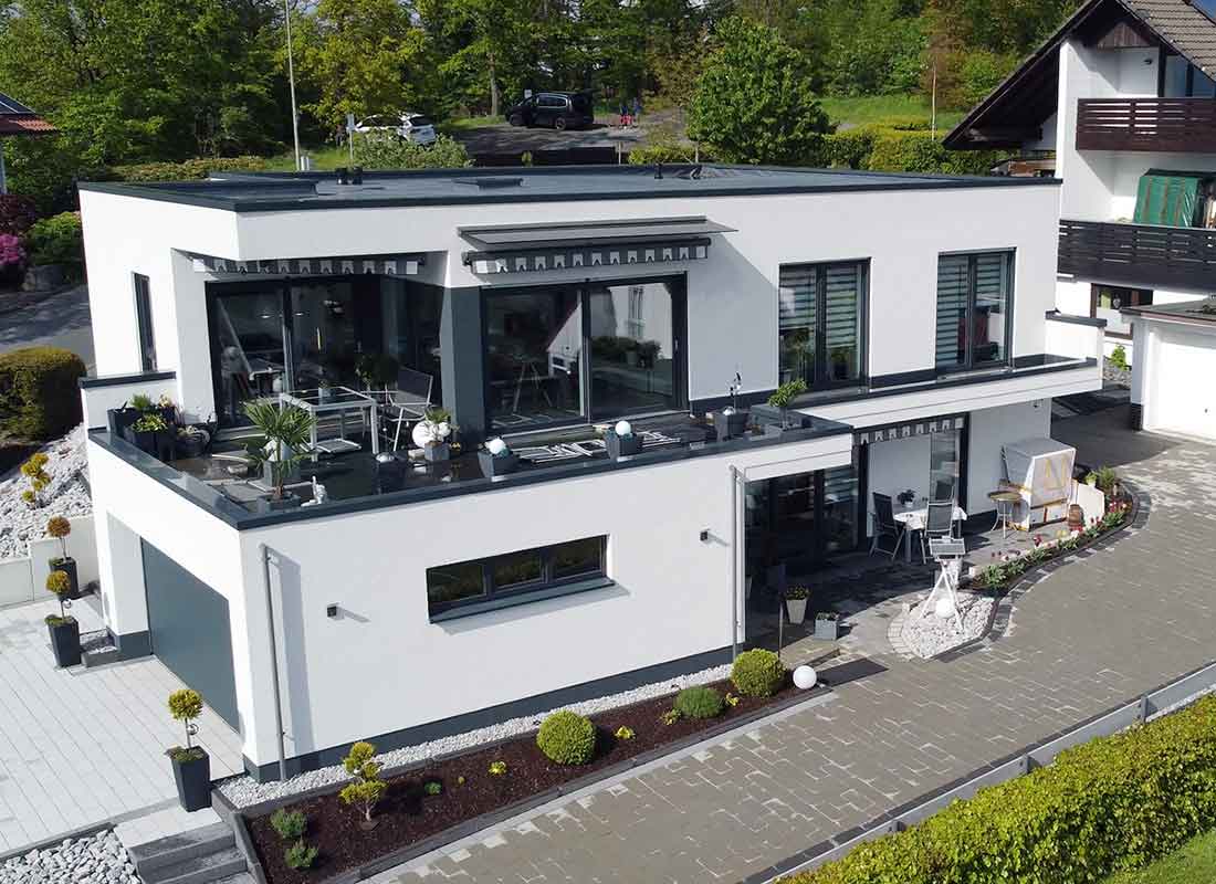 You can see a new building with a flat roof, a terrace on the first floor as well as a garage on the left side and another driveway in the foreground. In the background, you can see a small forest.