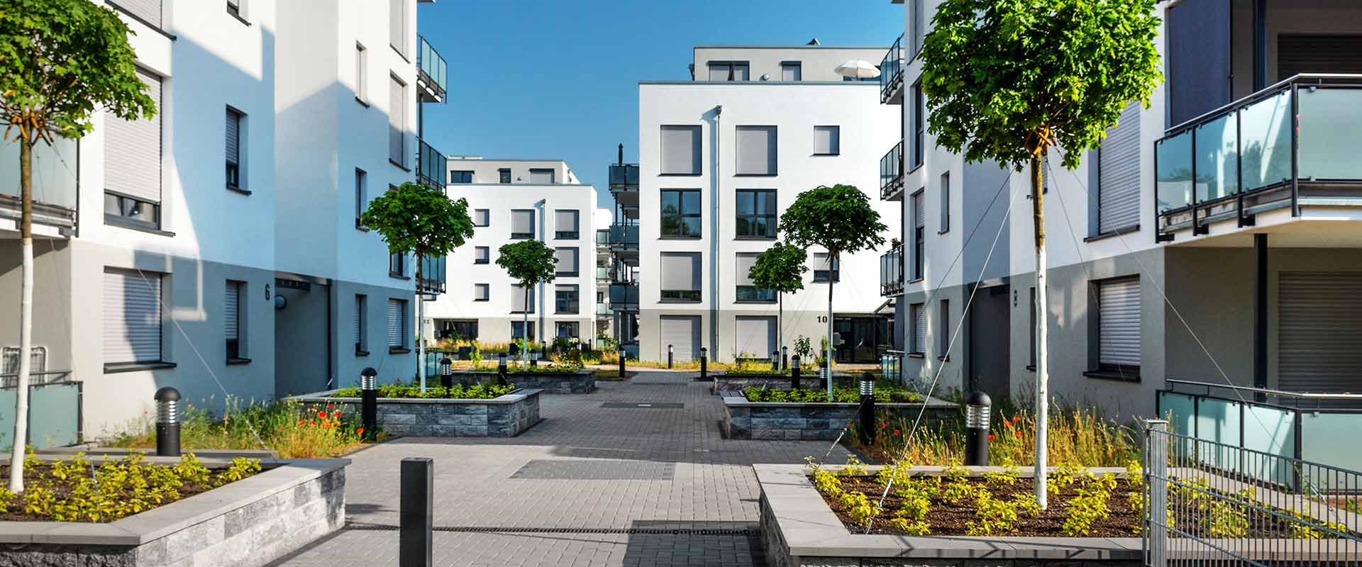 There is a footpath with planted areas and flower beds surrounded by multilevel residential buildings with flat roofs on both sides. In the background, more new buildings with flat roofs can be seen.