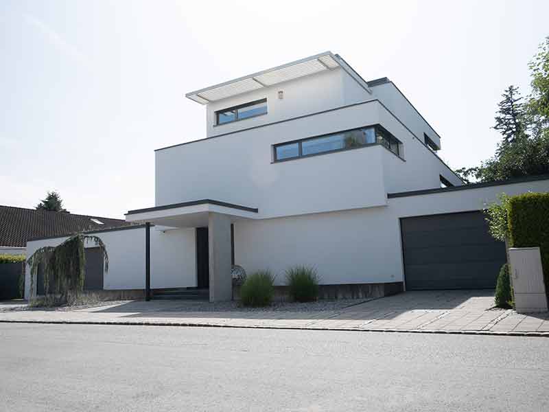 A two-story new building with a flat roof is illuminated by the sun from above with decorative gravel and small bushes in front of the front door, on the right you can see a closed garage door. In the foreground, you can see a street. 