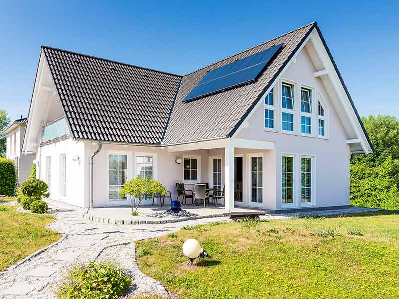 A family house with photovoltaics on the roof, a garden and a terrace in the foreground was photographed with a cloudless sky. In the background, you can see bushes and another house.