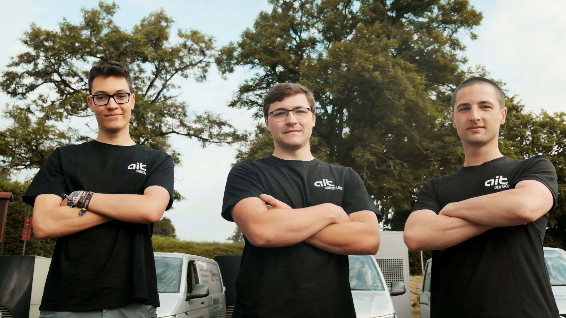 Three young men stand with their arms crossed and look at the camera with a serious expression. In the background, there are ait service buses, with heat pumps behind them. Further back, some green meadow and trees can be seen.