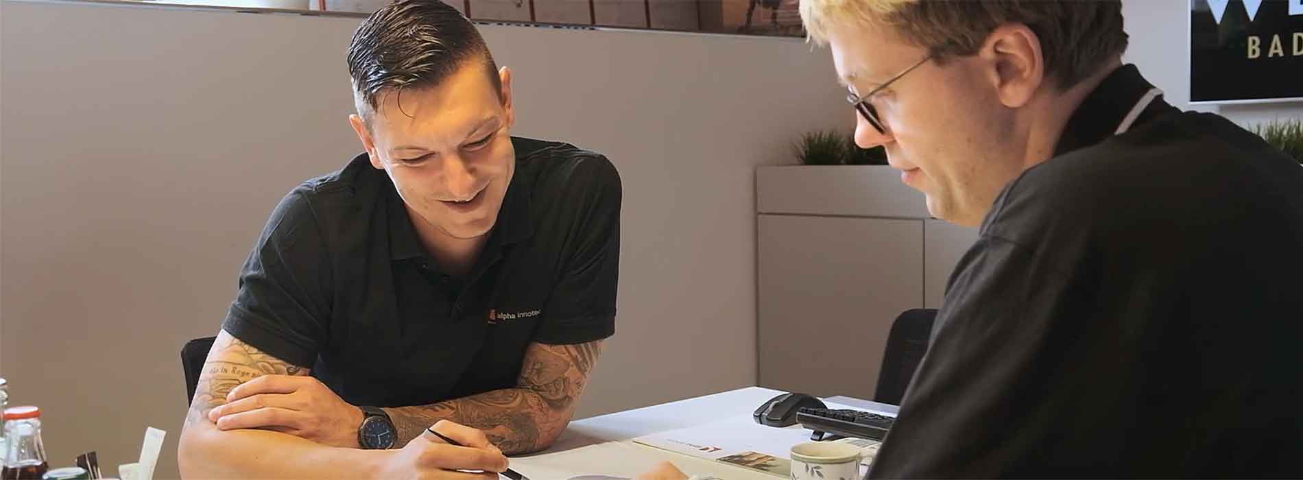A man with tattoos on his arm explains something to a customer at the table. With a pen, he points to paper sheets lying there, at which both of them are looking.