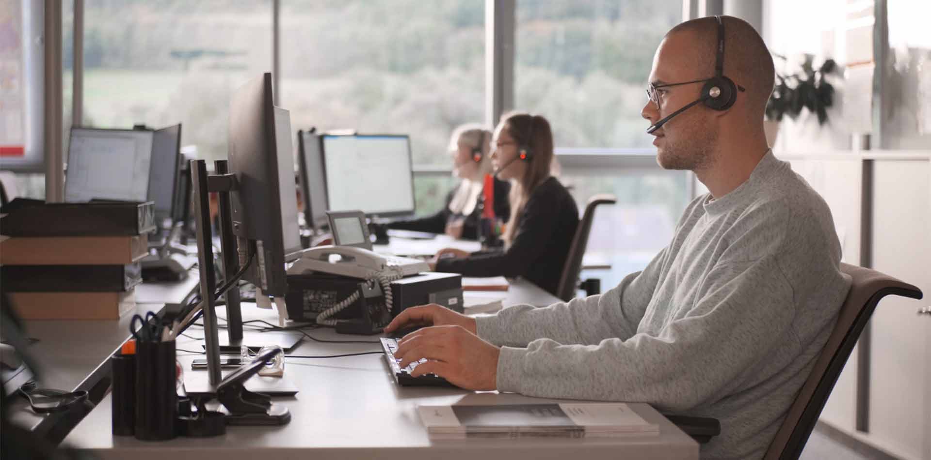 A man, photographed from the left side, is sitting in an office with a headset in front of PC screens and typing on the keyboard. In the background are two colleagues sitting next to him, also with their own PC and headsets on their heads.