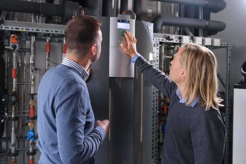 A man with shoulder-length hair stands right from a heat pump and adjusts something on the controller and display of a heat pump, while another man watches him from the left and looks at the heat pump. It is connected to many pipes in the background.