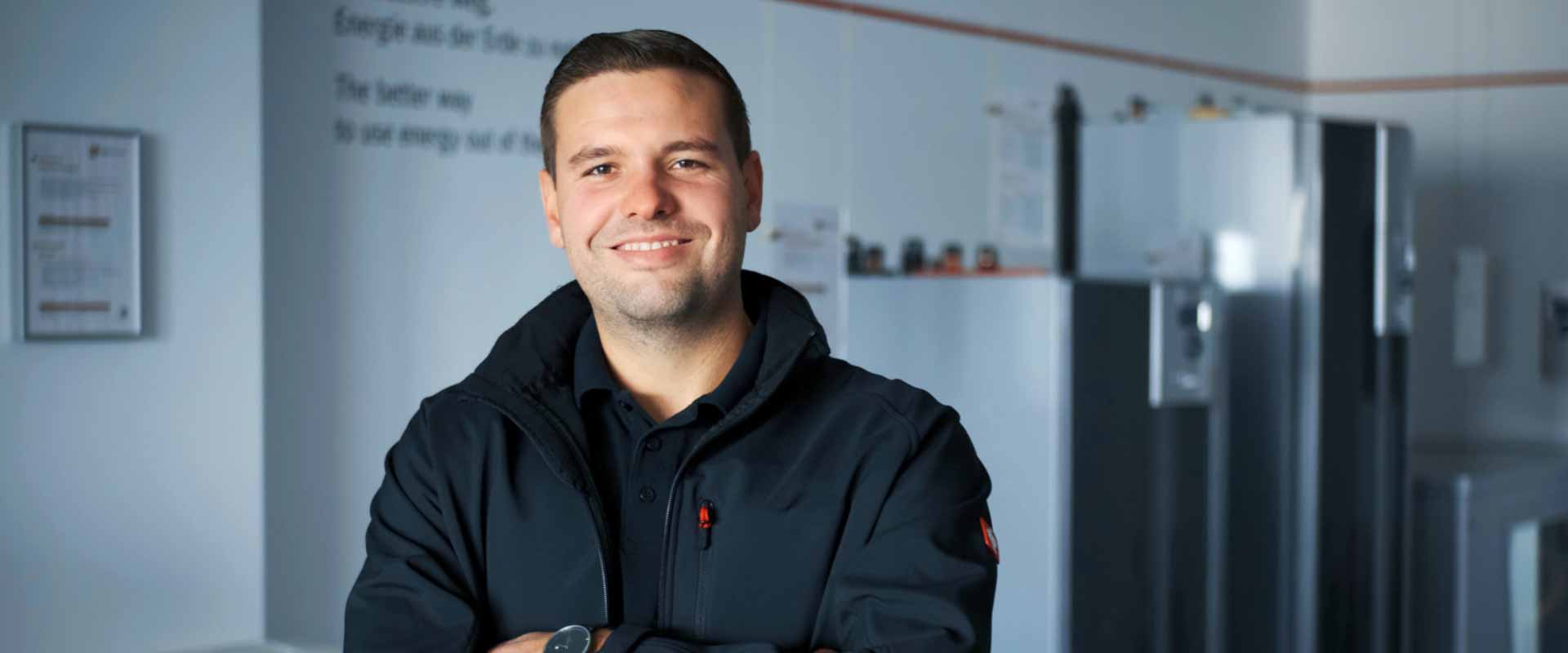 A man stands smiling, arms crossed, in front of several models of heat pumps that can be seen in the background.