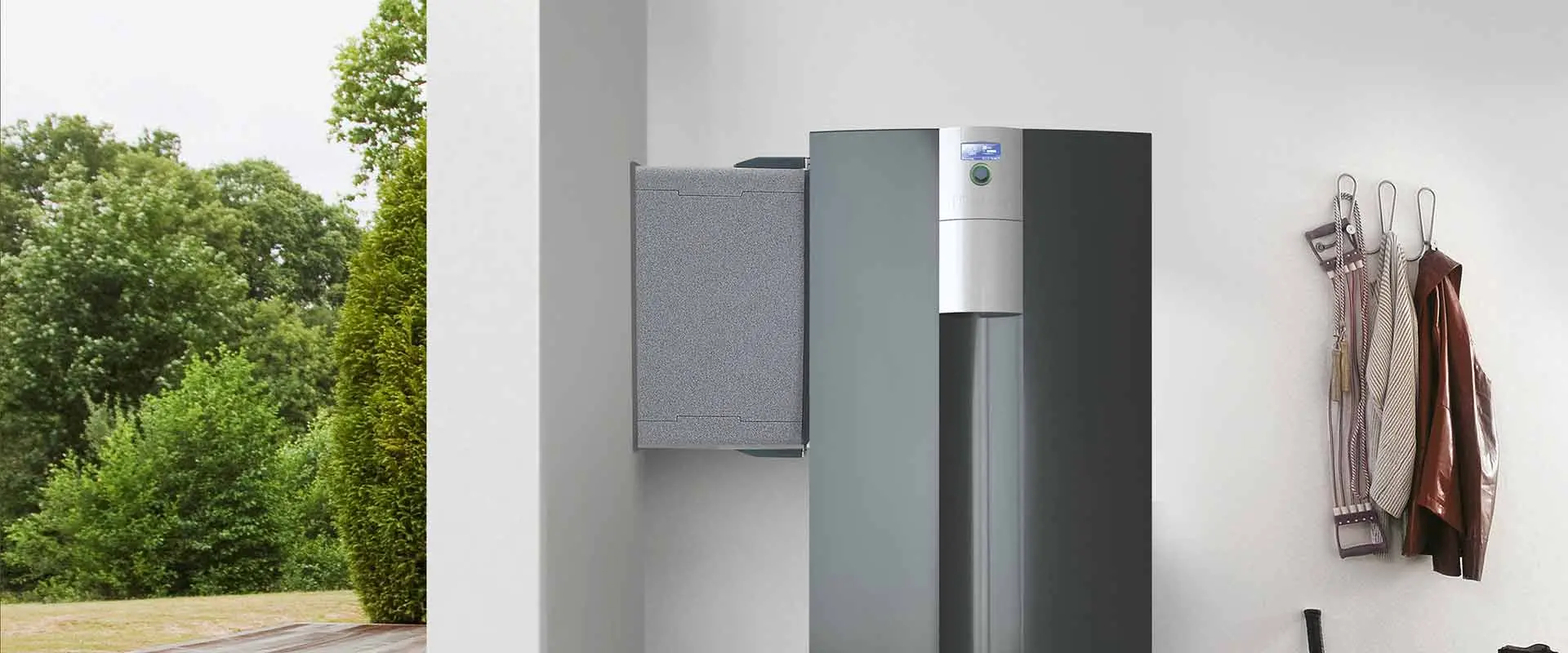 A cross-section of a room shows the air/water-heat pump LWCV indoors with an installed air duct extending into the wallon its left side. On its right there is a wardrobe and outside the left wall, there is some meadow and some trees.