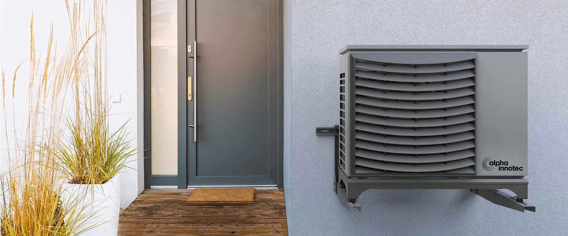 The heat pump Jersey is installed on a house wall and right next to the front door. On the left side of the door there are planted grasses.