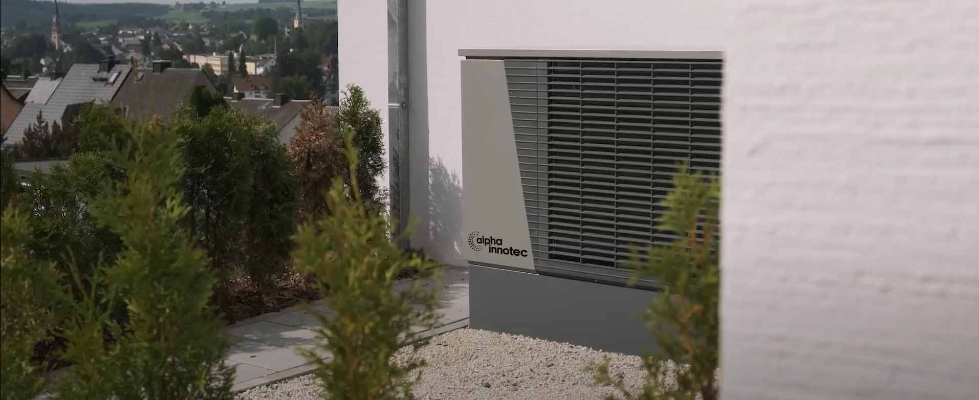 The heat pump LWD stands in front of a house wall on white decorative gravel, to the right of a paved path and surrounded by small bushes.
