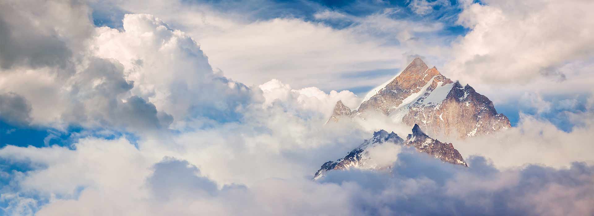 Snow-capped mountain peaks point through a cloud cover. In the background, you can see further cloudy sky.