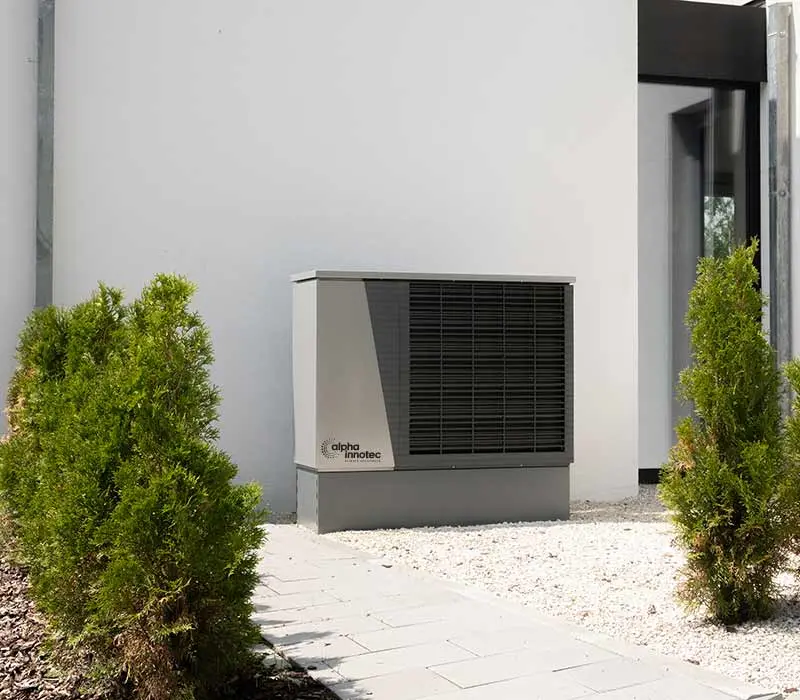 The air/water heat pump LWD is installed outdoors on white decorative stones in front of a house wall, next to a paved way and two small bushes.