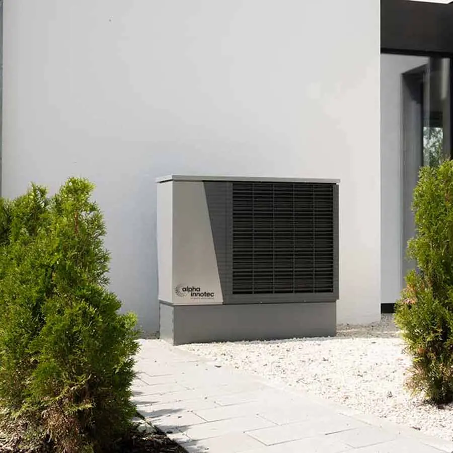 The air/water heat pump LWD is installed outdoors on white decorative stones in front of a house wall, next to a paved way and two small bushes.