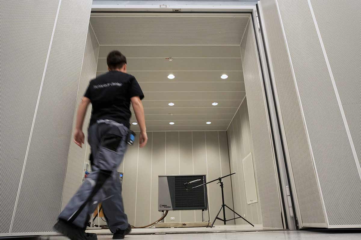A person is walking into an acoustic chamber and a heat pump can be seen further in the middle of the chamber with a microphone.