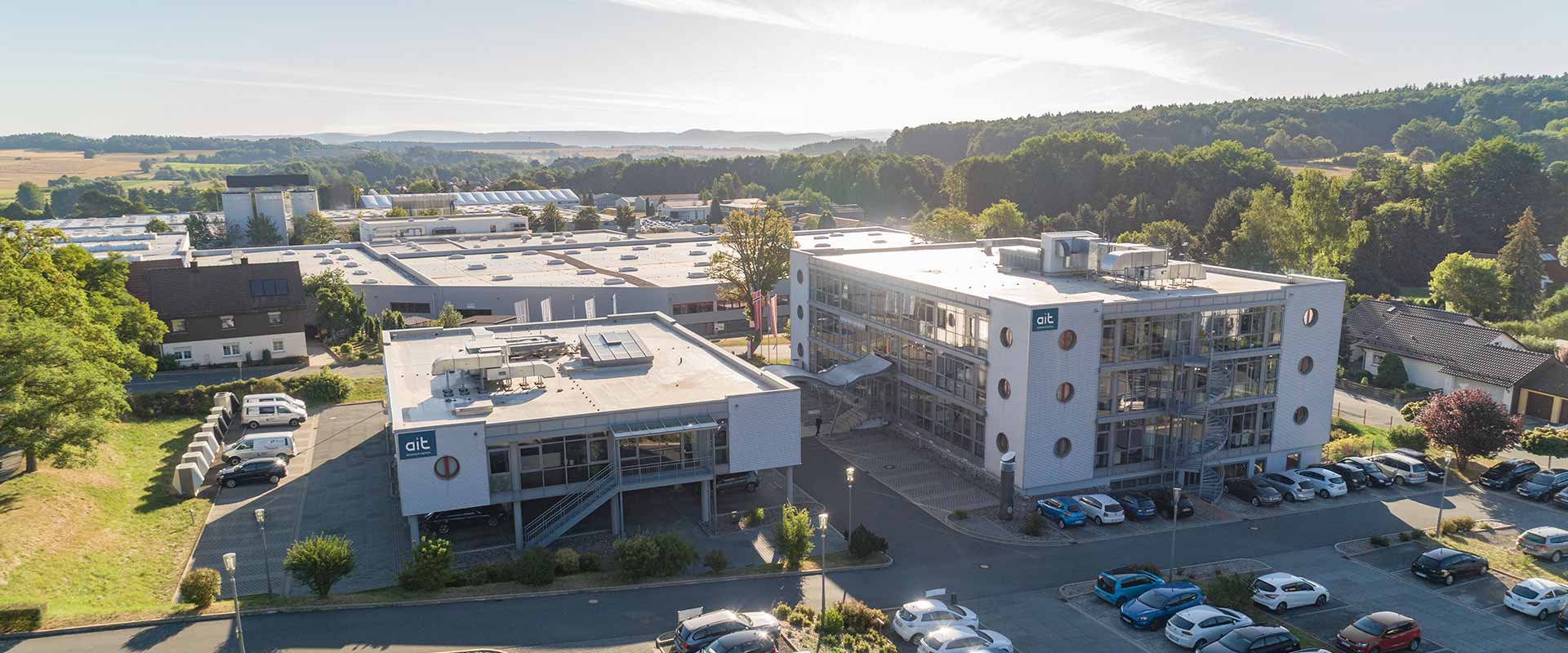 The company building is shown from an aerial view, illuminated by the sun, alongside the ait-Academy and the factory building in the background, which is mostly surrounded by forest.