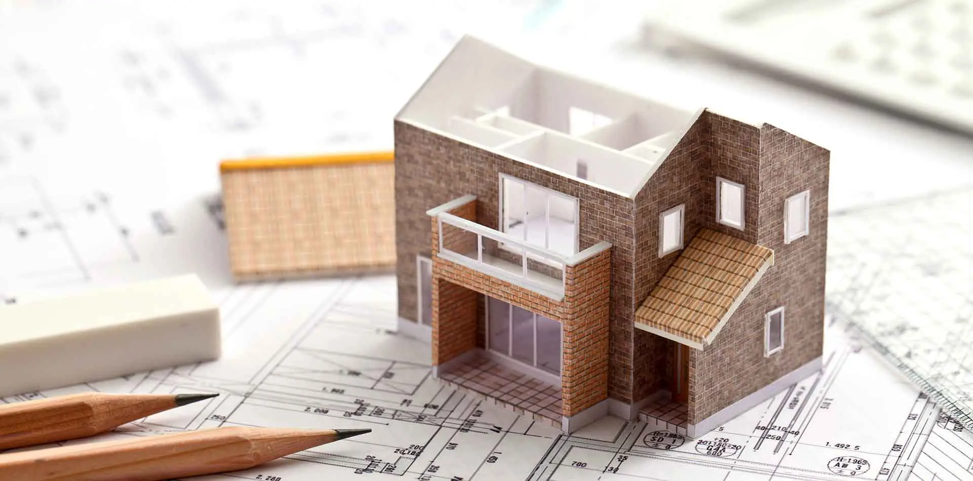 A miniature house is in the middle of the construction plan of a real house, with pencils and erasers on its left side.