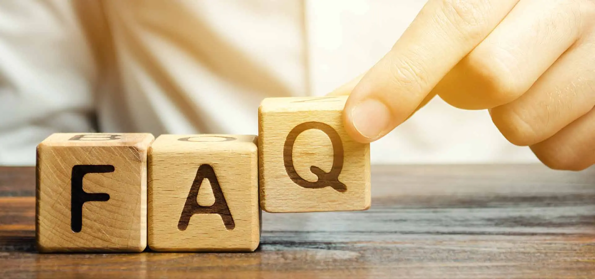 There are three close-up cubes made out of wood with the letters F,A and Q, one letter each cube. A person puts the last cube with the letter Q down to spell the word FAQ. 