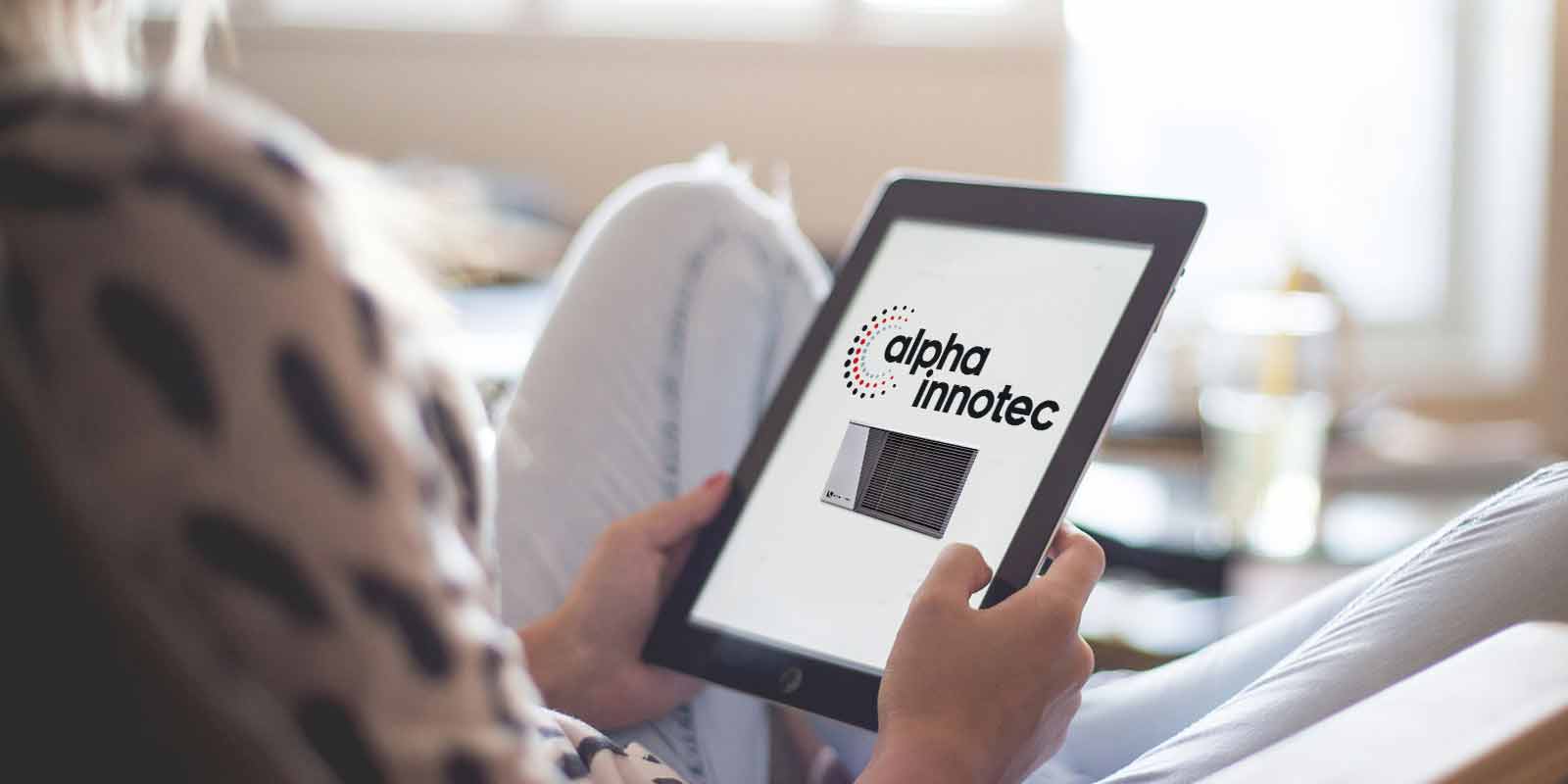 A young woman is sitting cross-legged with a tablet in her hands and on the screen there is the alpha innotec logo and a heat pump.