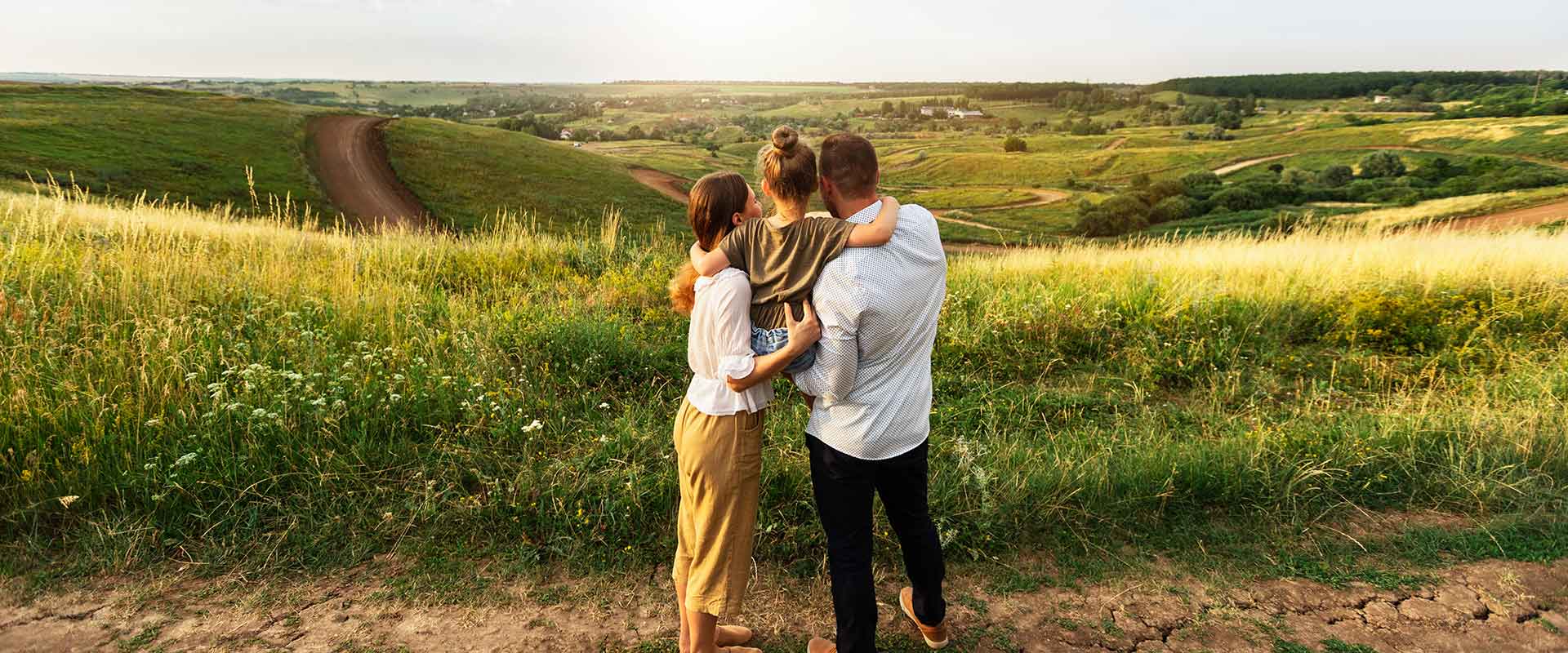 A couple is holding its daughter in its arms and all three of them enjoy a wide view over grassland and fields with some houses far away in the background.