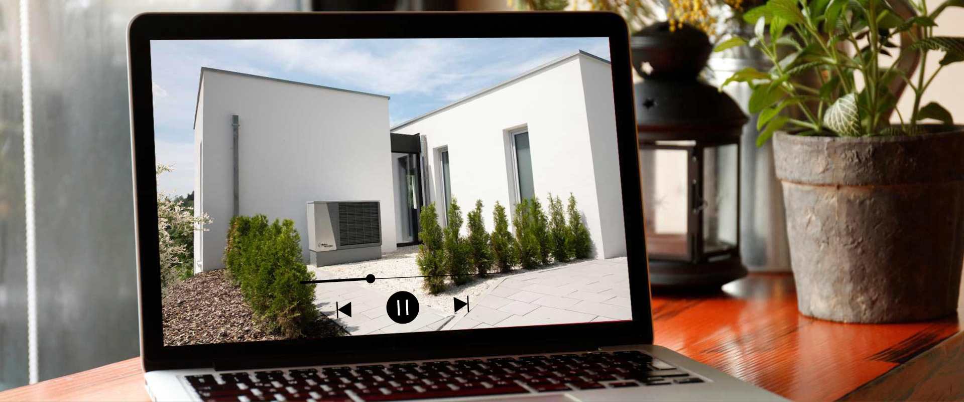 On a table is a laptop with a product video by alpha innotec playing on the screen and a potted plant on its right side.