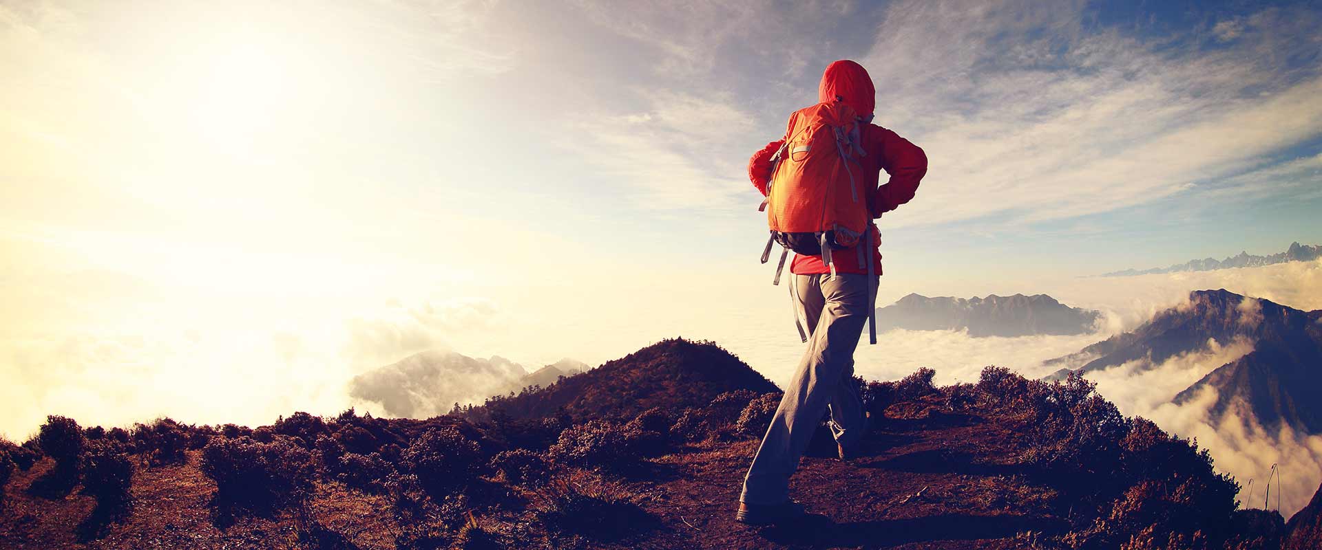 A woman with a backpack walks on a hiking trail on the top of a mountain while the sun shines on her from the left and clouds surround the tops of the other mountains in the background.